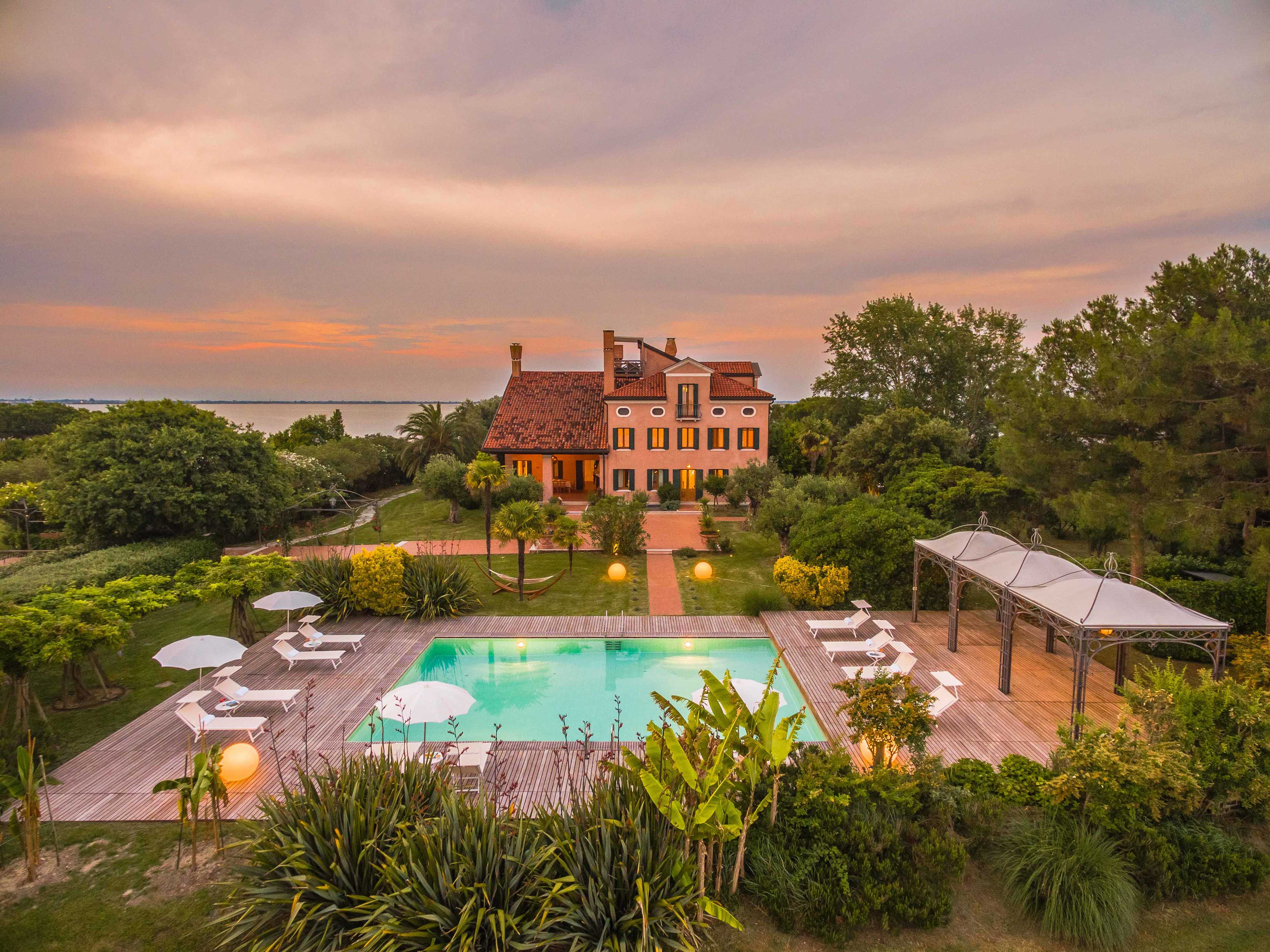 Wide shot of the villa and the swimming pool from the gardens. The square shaped aquamarine swimming pool is surrounded by sun loungers with the terracotta coloured villa in the distance. The sky is awash with hues of orange and pink.