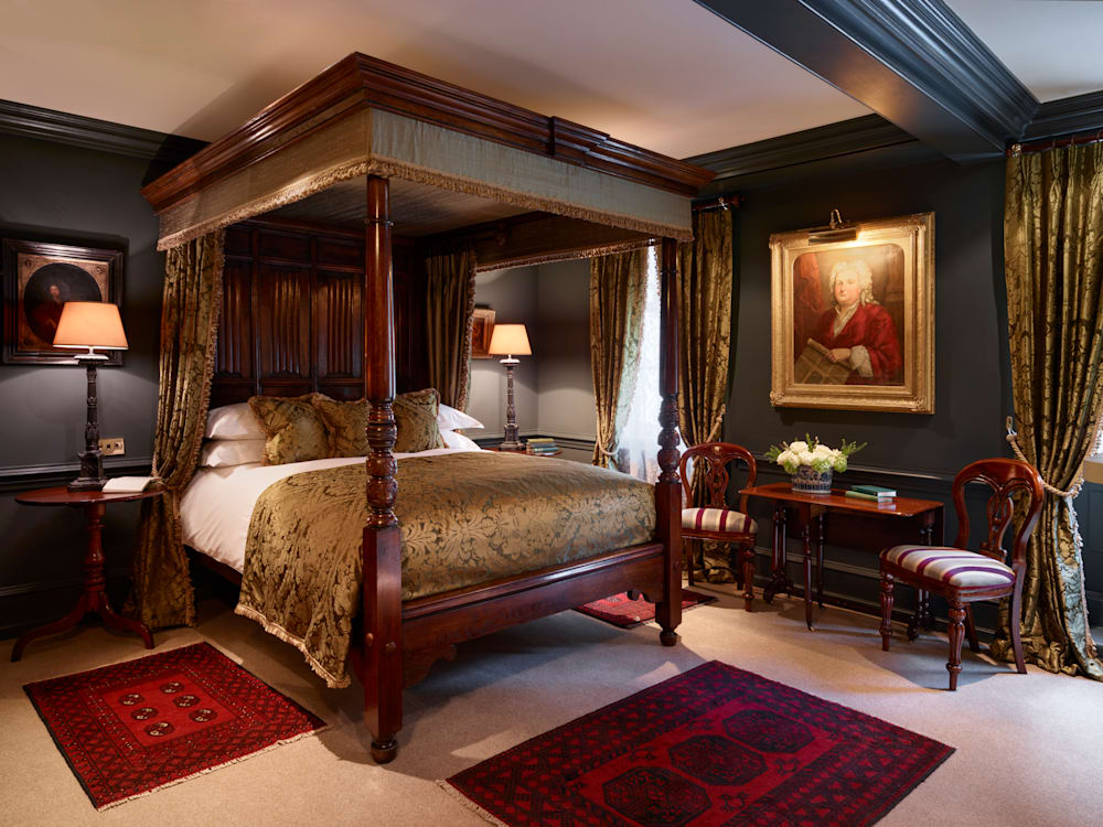 Ornate four poster bed in a richly decorated room. Deep red rugs with heavily patterned fabric curtains and deep grey/blue walls furnish the room along with oil paintings on two of the walls. 