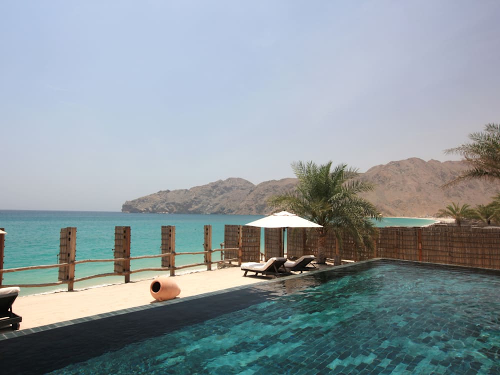 Private swimming pool and sun beds overlooking the beach and the aquamarine ocean. The craggy mountains are in the background reaching up to a hazy blue sky.
