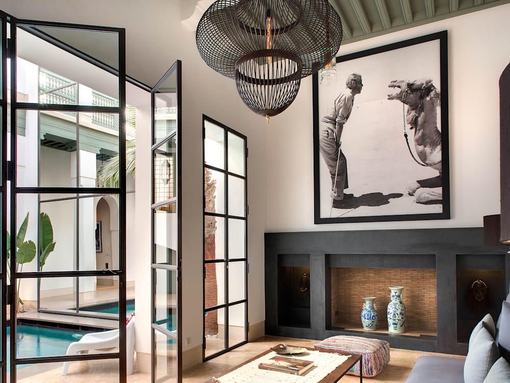 Photograph looking into the lounge with high ceilings, coffee table and sofa. There is a hollowed out fireplace with ornate vases. Above the fireplace sits a metre high black and white artwork of a woman and a camel. There are floor to ceiling windows looking out onto the swimming pool and courtyard.