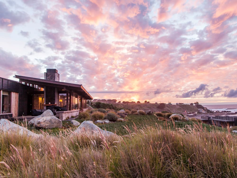 Hotel lodge overlooking the sea. Wild grasses frame the foreground and there is an orange, pink and purple watercoloured sunset scattering the fluffy clouds across the sky