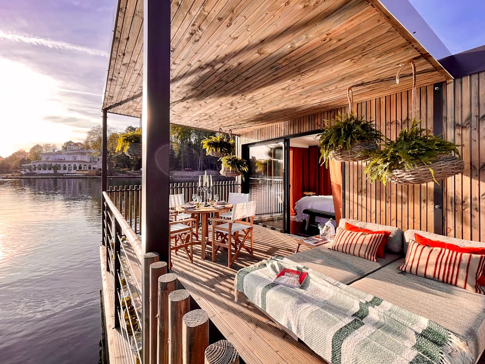 Sun loungers and terrace above the lake. The sun is setting and the sky has deep hues of purple, orange and blue. A table is set on the terrace for dinner and there is a glimpse of the bedroom in the open door. 