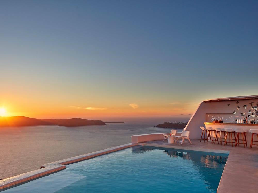 Deep orange sunset over the sea, the golden light hitting the terrace and swimming pool. The bar is in the far corner with two chairs set up overlooking the sea.