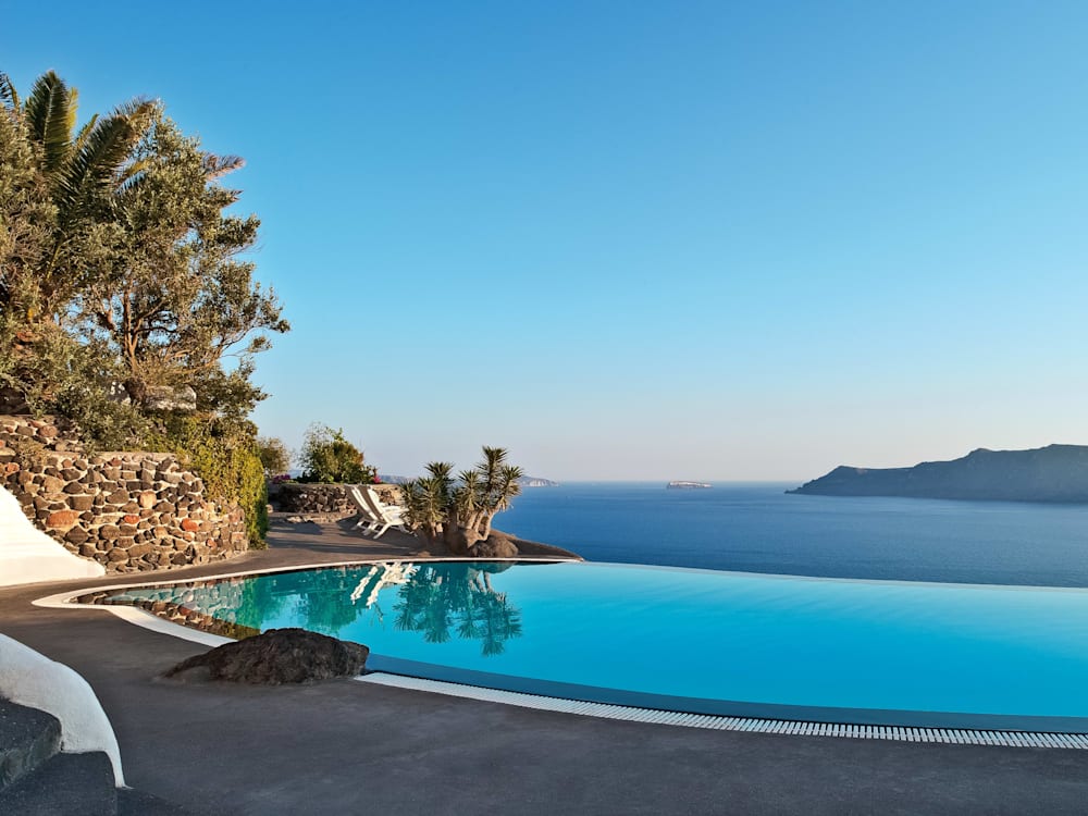 Infinity swimming pool overlooking the ocean. Trees frame the edge of the swimming pool and two deck chairs have pride of place on the edge of the cliffs. The late afternoon sun is illuminating the terrace, golden hues dominating the scene. 