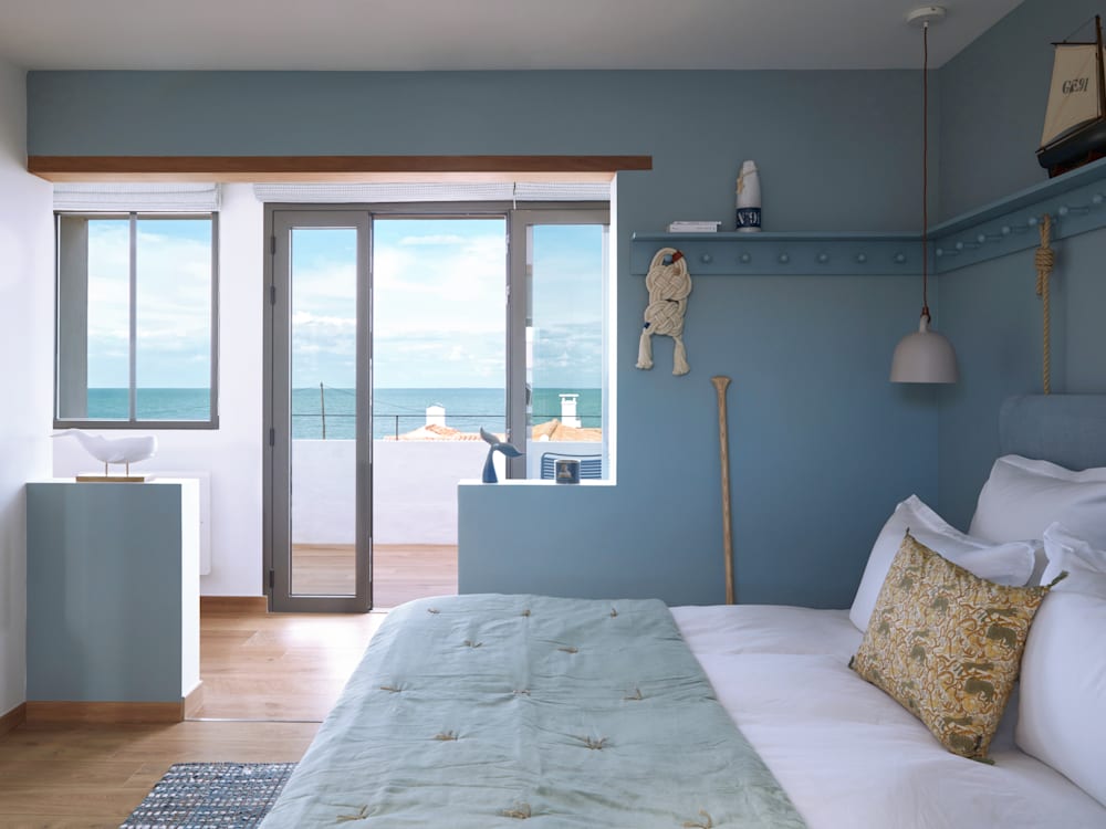 Tranquil bedroom with calming blue walls looking out onto a terrace with the ocean in the distance.