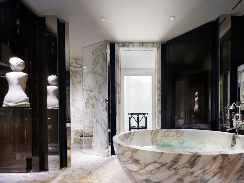 Oval shaped marble bath tub in a black and white marble bathroom. A Roman bust female statue is on the side of the room. 