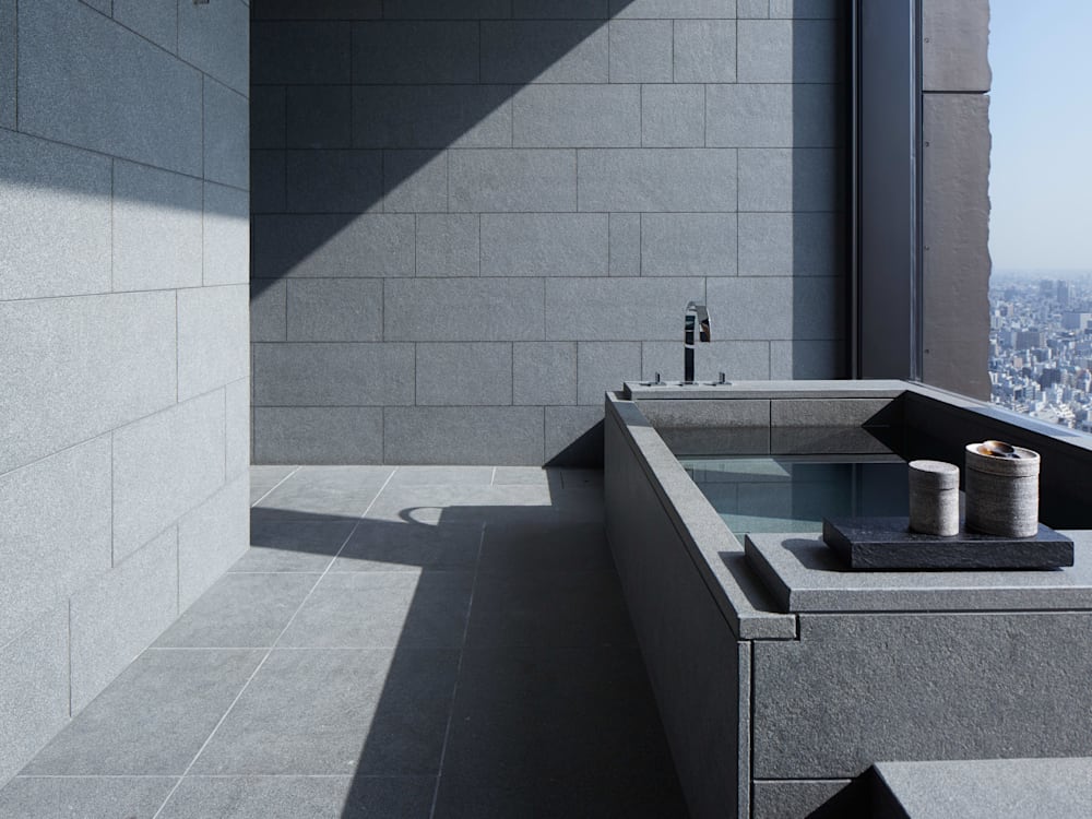 Minimalist slate grey bathroom. The bath is the only thing in the room next to the floor to ceiling window looking out onto the city far below