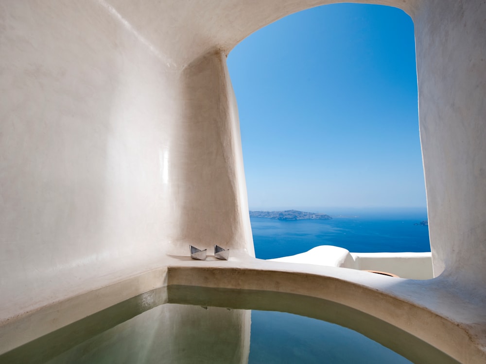 White walled room with a huge open window. The bath is looking out over the ocean. The sky is brilliant blue melting into the deep blue sea.