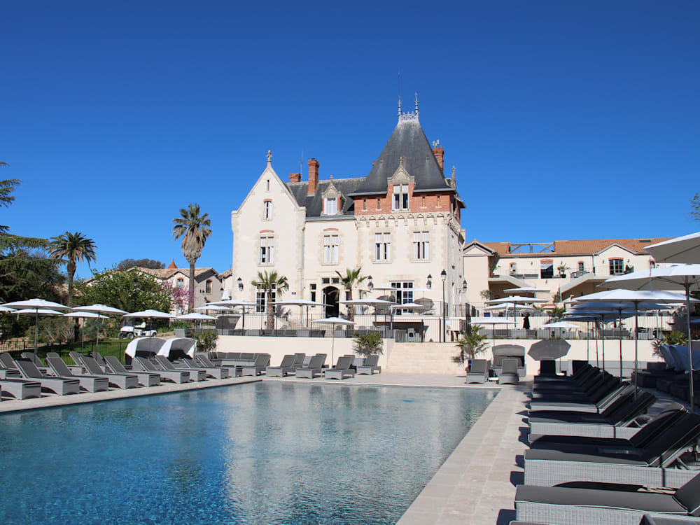View of the chateau from the swimming pool. Sun loungers and parasols surround the pool in front of the white building of the chateau. The deep blue sky doesn't have a cloud in sight. 