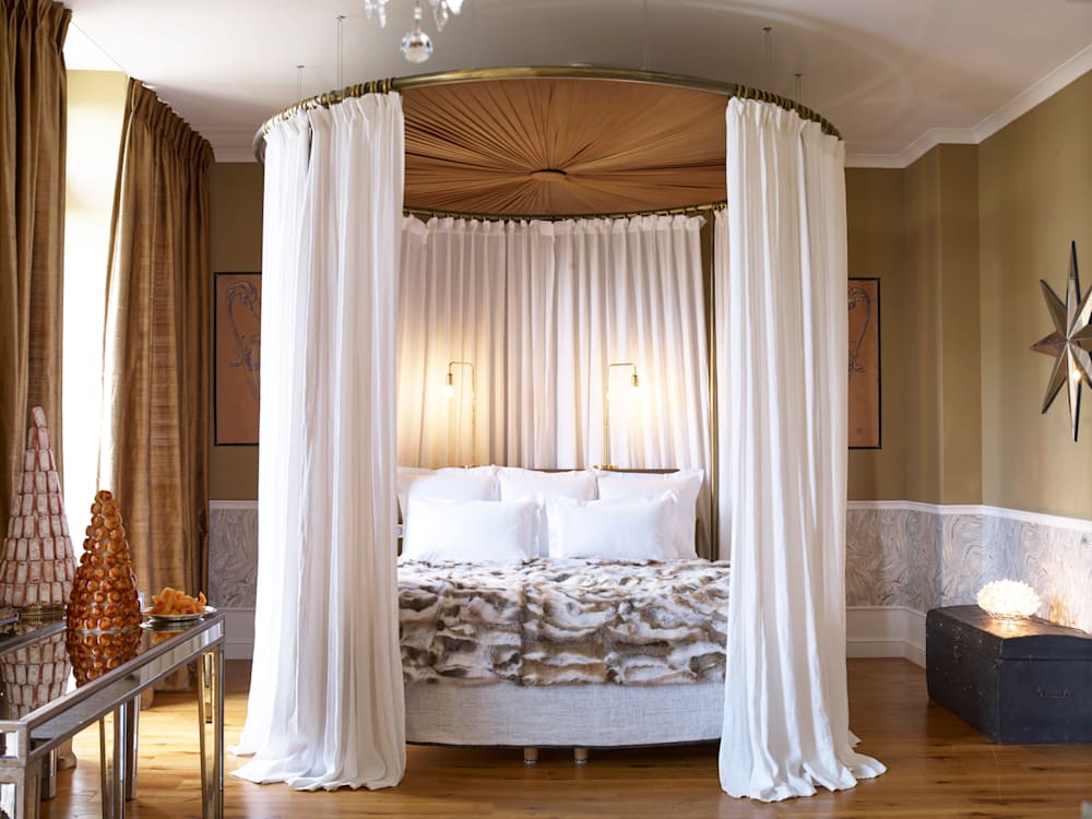 Grand four poster bed with sweeping white floor to ceiling curtains surrounding it. The light is streaming in through the windows into the tranquil setting and warm golden hues of the room. 