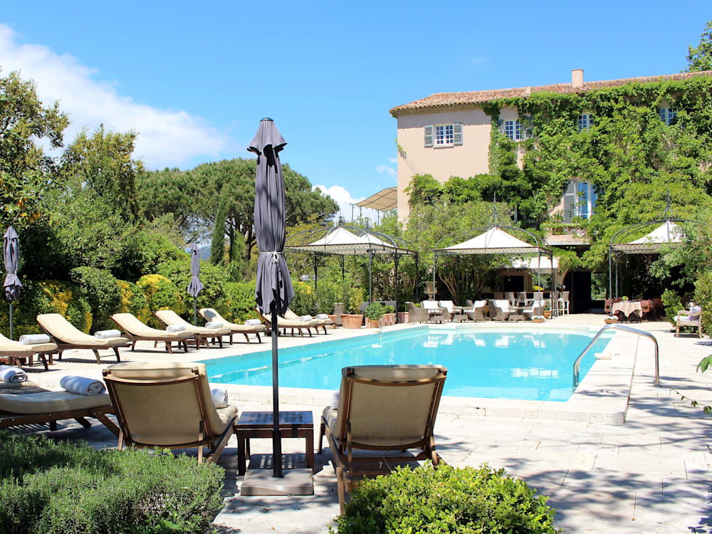 Photograph of the rectangular swimming pool surrounded by sun beds. The ivy covered building of the hotel is in the background against bright clear blue skies. 