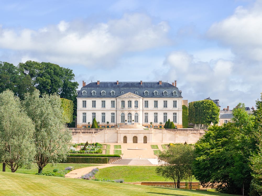 View of the chateau from the gardens. It is a white building with a grey/blue roof. There is a grand staircase leading down into the sweeping gardens. Fluffy white clouds are scattered across the blue sky.