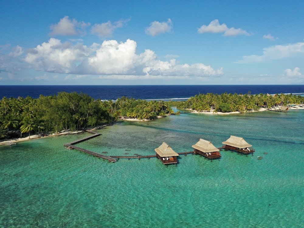 Aerial view of the hotel, the rooms on the lagoon with a walkway leading to the island. The deep blue of the ocean stretches out into the distance.