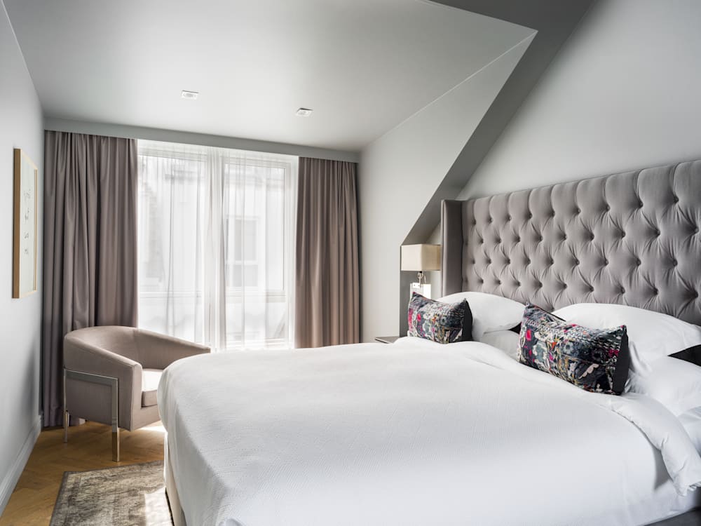 Huge King sized bed with plush white bedding at Sandhotel, Iceland. There is a chair in the corner in muted hues that are in line with the curtains and headboard. Floor to ceiling windows are at the end of the room with light streaming in. 
