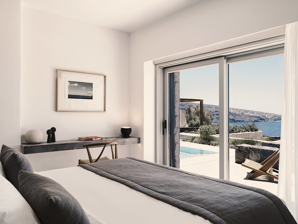 View from the bedroom. Big double big fills the frame in the foreground, there is a table and chair in the corner of the room with an artwork hanging on the wall. French doors look out onto a terrace with a pool on the edge of the cliff. Bright blue skies and the deep blue sea stretches out into the distance