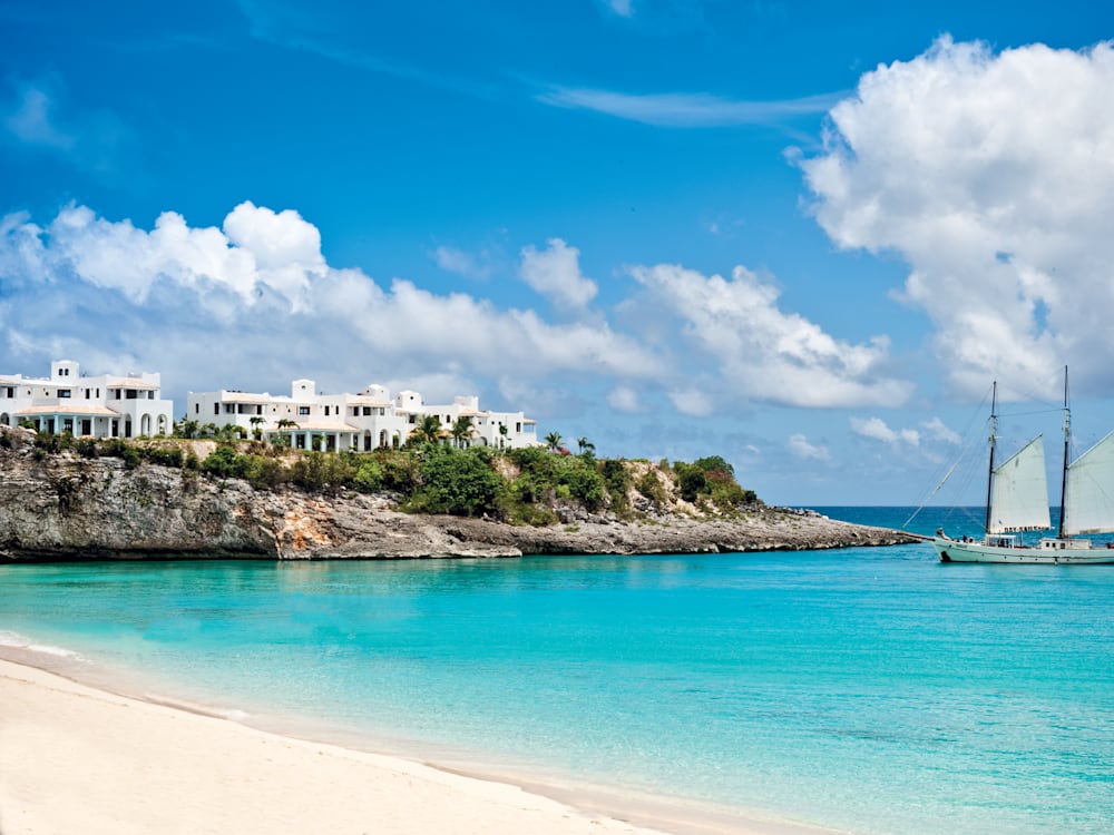 View of the white hotel on the cliff tops from the white sandy beach, there is a sailing boat resting on the azure blue waters