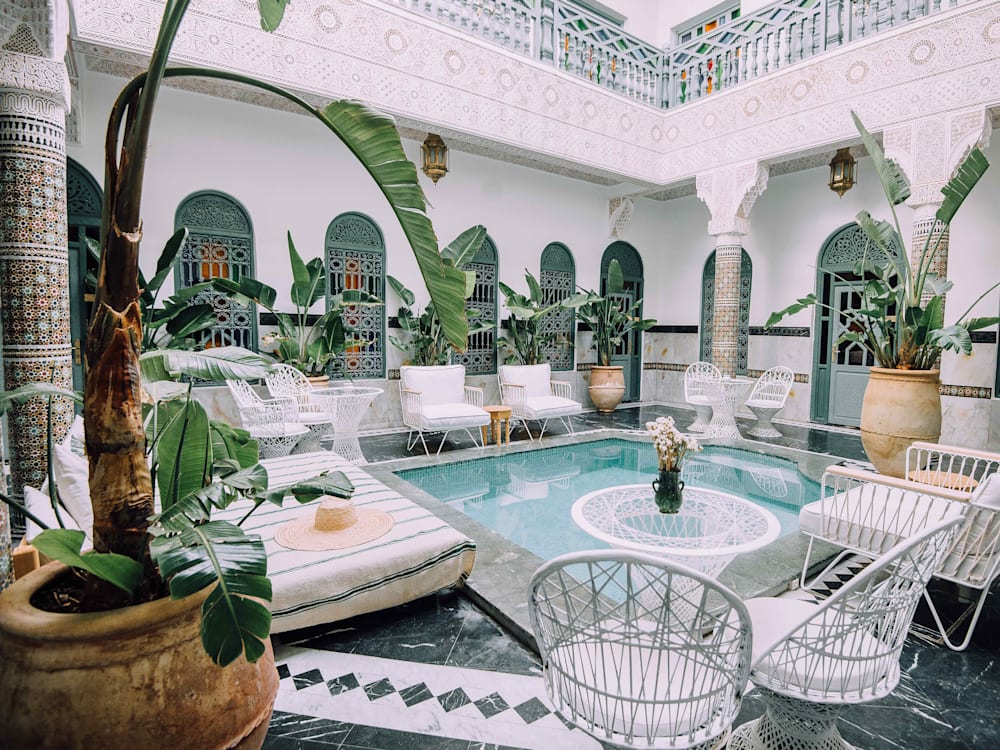 The courtyard patio with chairs and sun loungers surrounding a square bright blue pool in the centre. The walls are tiled in colours of green and blue with bright white pillars. 