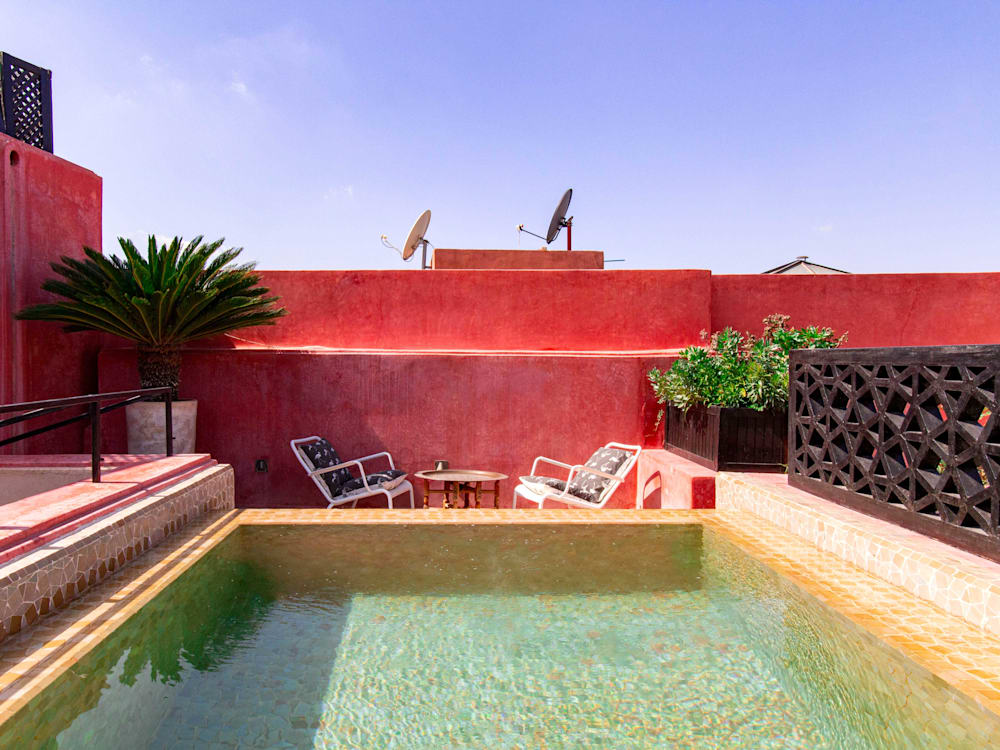 The aquamarine rooftop swimming pool is in the foreground with two chairs and a table behind it. The pool is framed with the bright red building and standing pot plants with clear blue skies overhead