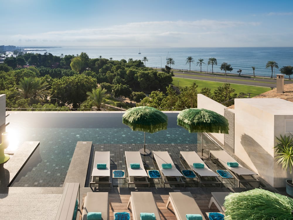 Rooftop swimming poor with the sun bouncing off the clear water. Sun loungers have pride of place next to the swimming pool. The infinity rooftop pool looks out over the trees and the ocean and palm trees are in the distance. Bright blue skies overhead.