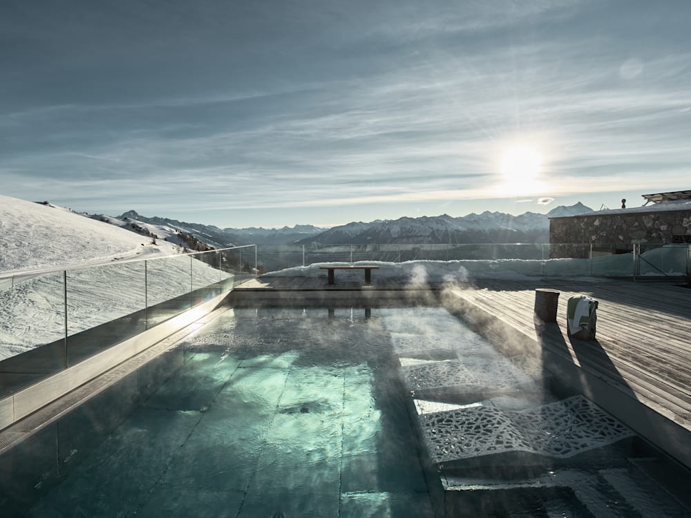 Rooftop swimming pool overlooking the mountains. The sun is high in the sky, reflected in the aquamarine swimming pool. Snow is ontop of the roof and steam is coming off the heated pool. 