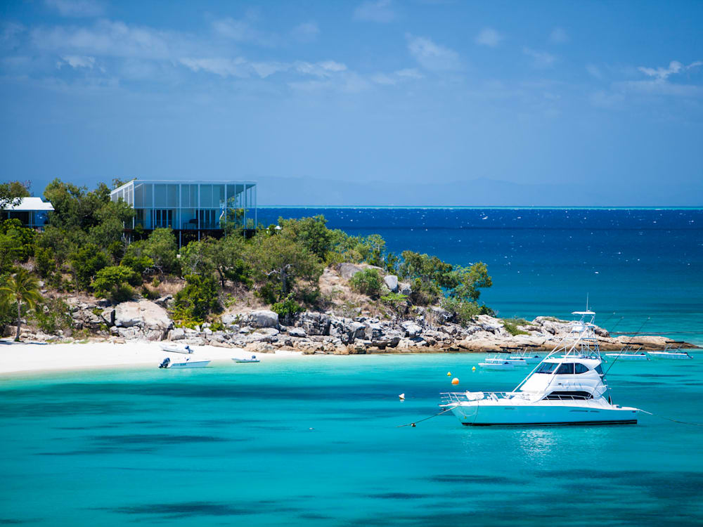 View of the hotel from the sea, the sandy peninsula has white sands and the hotel is at the top of the hill. There is a boat in the bay on top of the crystal clear waters. 